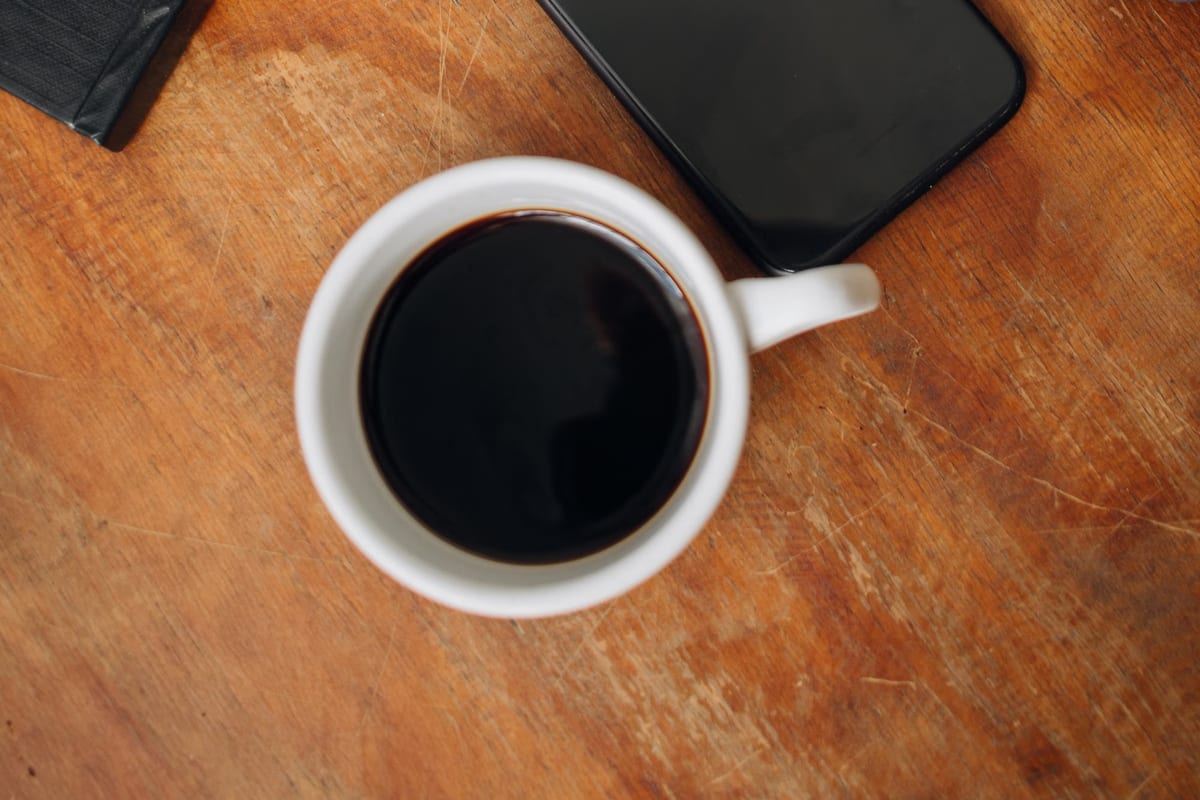 A cup of black coffee and a phone on a wooden table, photographed from above