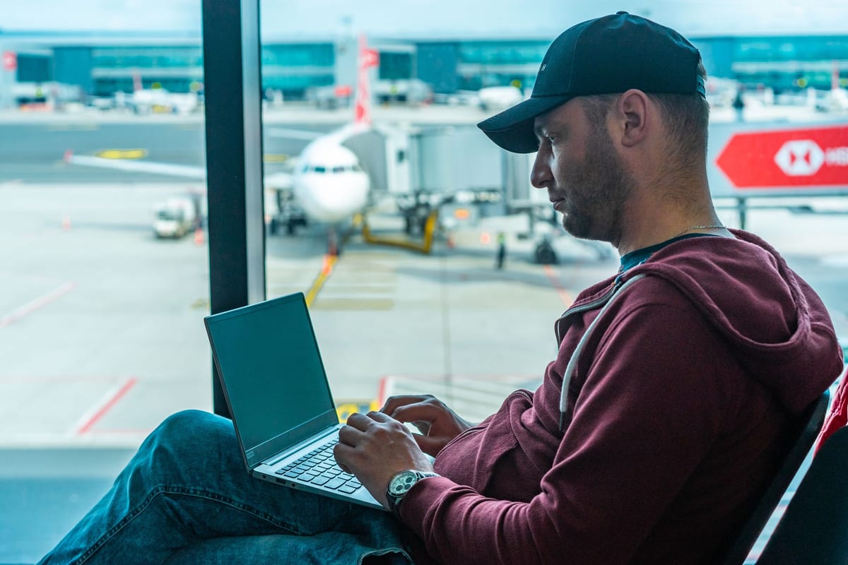 A man in a cap working on a laptop at an airport gate, with an aircraft outside the window