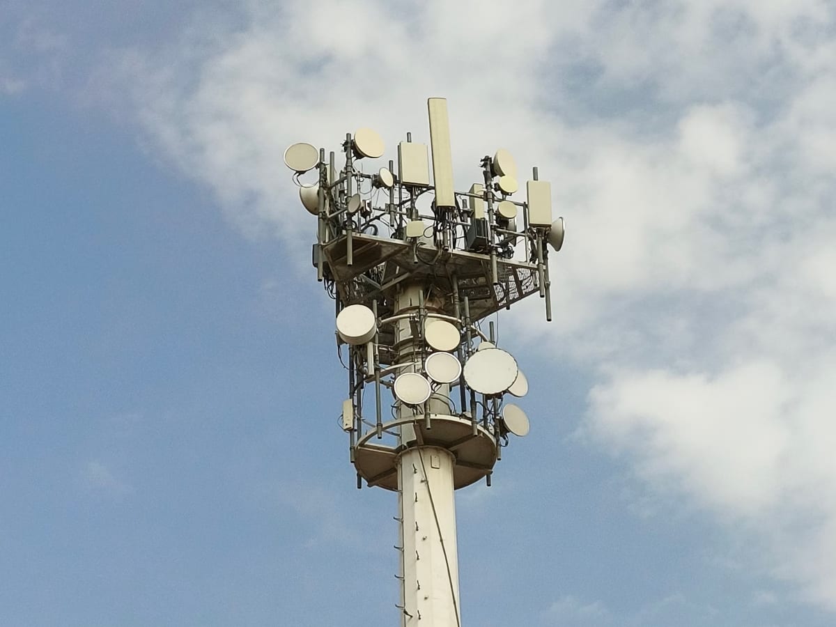 A mobile network mast carrying antennas and microwave dishes against a cloudy sky