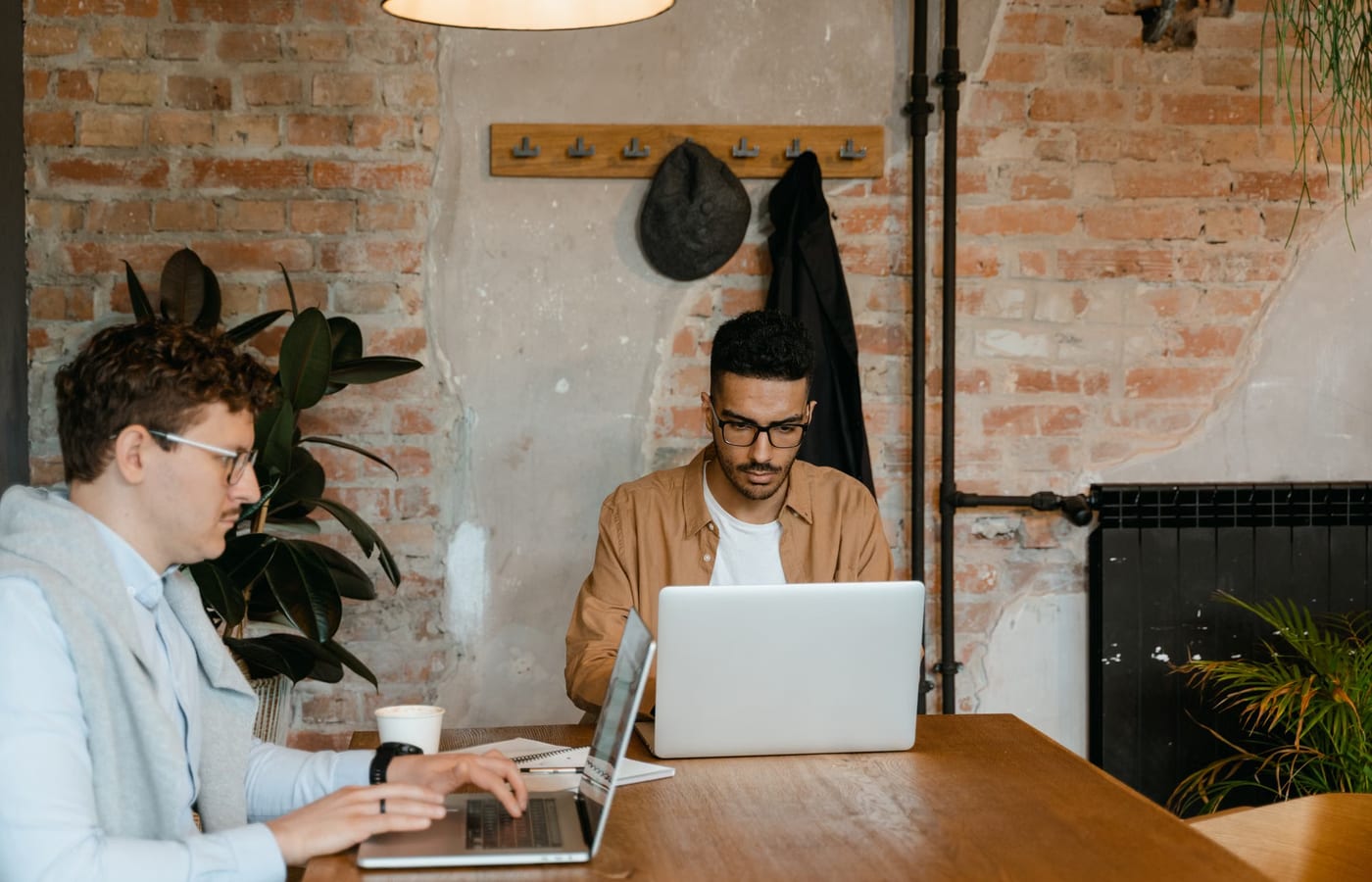 Two people working on laptops at a table in a brick-walled workspace